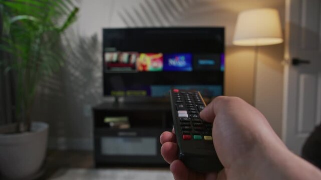 A man's hand skillfully maneuvers a remote control to switch channels on a smart TV, creating an engaging atmosphere in a cozy living room filled with modern d&eacute;cor