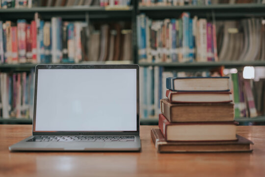 A simulated image of an empty laptop screen on a wooden table with a stack of books in a library, with a library as the backdrop.