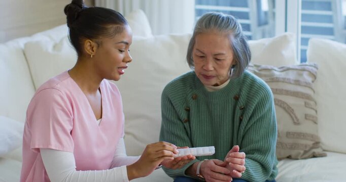 Diverse caregiver in scrubs showing pill pack at home, senior woman in green sweater learning doses