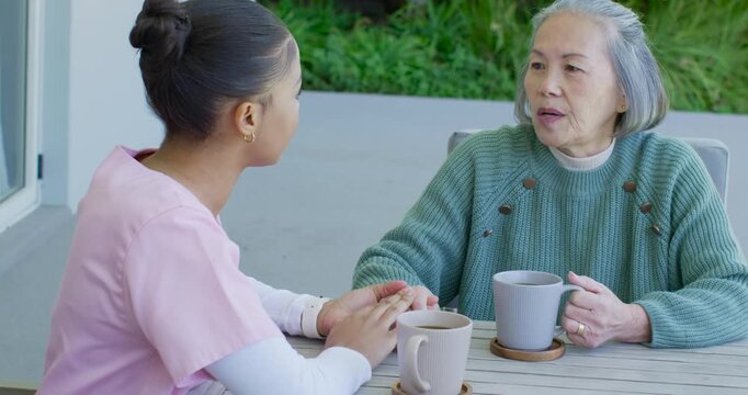 Female aide in pink scrubs, senior woman on patio holding hand, senior speaking for comfort, mugs