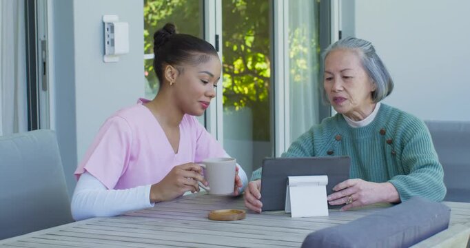 African American caregiver in scrubs holding mug guiding Asian Senior after tablet setup on patio