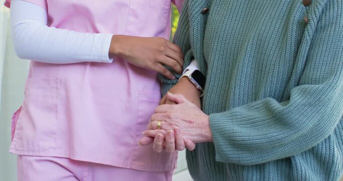 African aide in scrubs holding senior Asian woman after slip adjusting smartwatch talking by glass