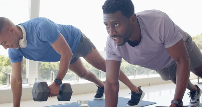 Asian and African American men starting plank, performing dumbbell rows near windows for core tone