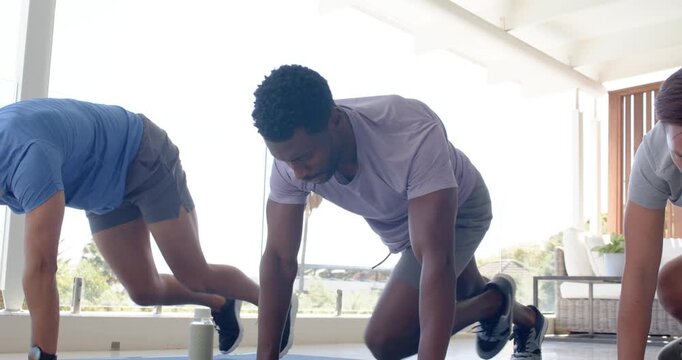 Diverse male trio starting plank, driving alternating knee tucks on blue mats on patio with bottle