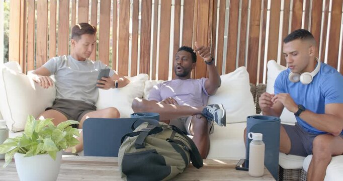 Diverse male friends relaxing while center man raising hand and speaking on patio sofa with phone