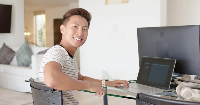 Mid Adult Asian male in striped tee typing on laptop at desk, noticing camera, pointing watch