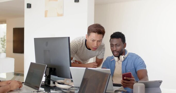 Diverse male coworkers working, showing red phone, leaning over laptops and monitor at glass table