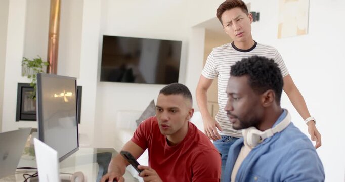 Diverse male coworkers walking in leaning over monitor and laptop sharing phones at glass desk