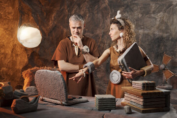 Caveman and Cavewoman Collaborating on Stone Laptop – Prehistoric Office Teamwork and Data...