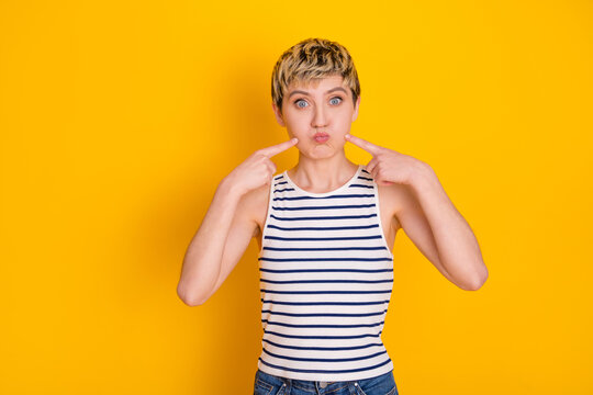 Young woman with short blonde hair wearing a striped tank top making a playful pout and pointing to her cheeks on a bright yellow background