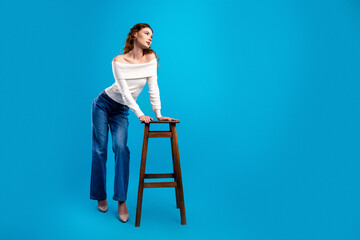 Fototapeta premium Fashionable woman in off shoulder white sweater and blue jeans posing elegantly with a wooden stool on a bright blue studio background