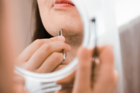 Reflection in a mirror of a woman using tweezers for facial hair removal on her chin, managing unwanted hair and hormonal changes in her home beauty routine.