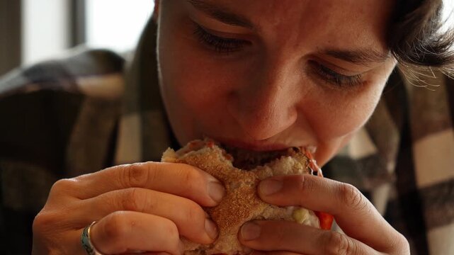 Young woman eating a fresh burger near window light in modern cafe interior. Concept of fast food lifestyle comfort hunger lunch and casual urban dining. Close up portrait