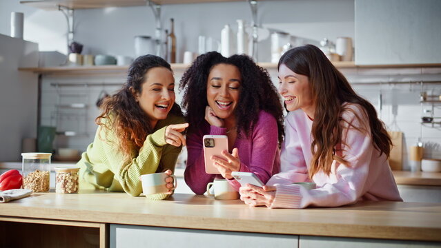 Multiracial women watching cellphones standing modern kitchen interior closeup