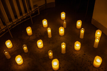 Overhead shot capturing a collection of lit candles placed randomly on a dark tiled floor. The scene is set in a dimly lit interior space, with the warm golden light of the candles providing the prima © Semiglass