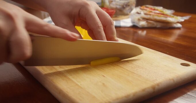 Close up of hands slicing fresh yellow bell pepper into thin strips with a kitchen knife on a wooden cutting board during vegetable preparation for home cooking.