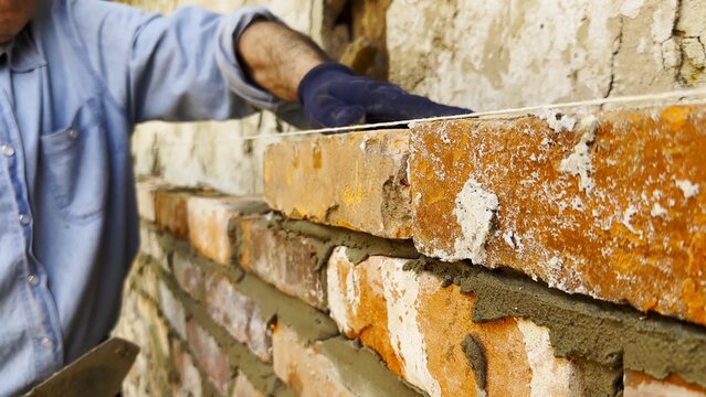 Adult male hands in protective gloves apply cement with a trowel and place bricks to build brick wall for house. The worker carefully spreads mortar and sets each brick during the construction process