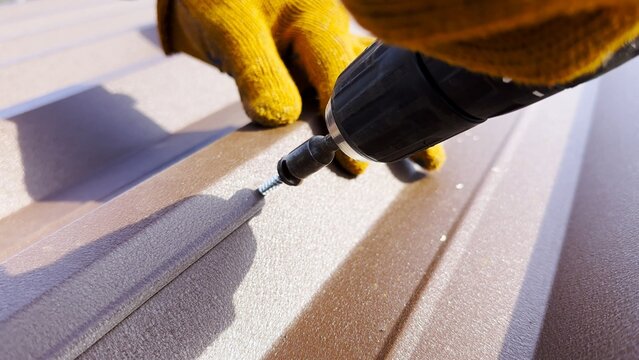 Male hands in gloves use cordless screwdriver to fasten roofing sheets in house roof. Young worker secures screws into surface performing careful installation work during residential construction