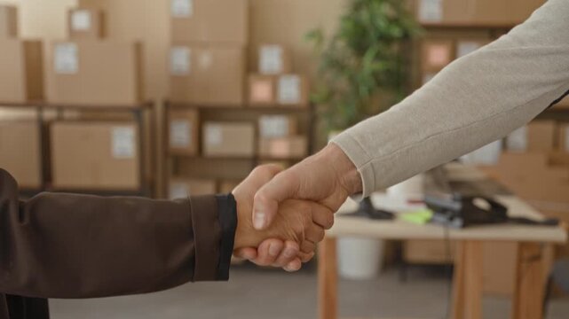 Man handshake with visible hands over stacked parcel boxes in warehouse shipping area, two middle age people sealing deal; trust partnership.