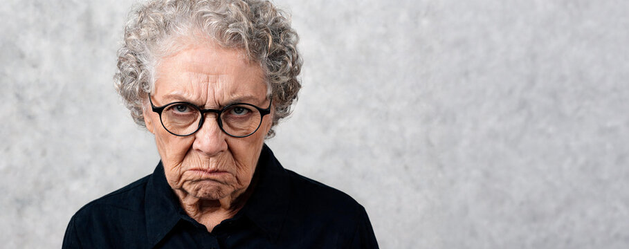 Angry casual senior woman with curly hair and glasses looking at camera. Portrait of displeased mature lady showing negative emotion. Human facial expression, discontent and mood concept.