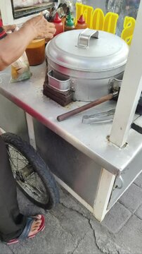 An Indonesian street food vendor preparing bakso (meatballs) and closing the metal steamer lid on his traditional food cart.
