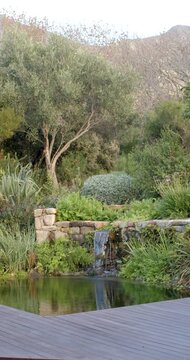 Vertical video: Gravity pulling cascade over stone lip in garden and stirring pond ripples