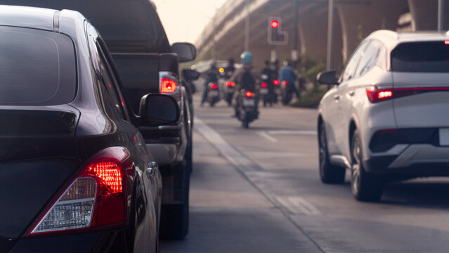 Abstract of black car and a white SUV move slowly through a congested urban avenue. Multiple delivery motorcycle riders can be seen navigating between lanes ahead in traffic. Overpass by the roadside.