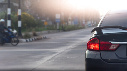 Fototapeta premium Rear corner close up of a dark car showing its illuminated red tail light. Car is stopped on a wide city road with soft daylight. Blurred of motorcycles waiting to cross the intersection.