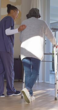 Vertical video: Nurse in scrubs guiding senior using walker hand on back practicing gait at home