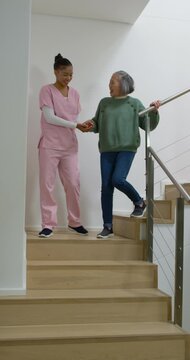 Vertical video: African American caregiver taking hand of Asian Senior woman helping descend stairs