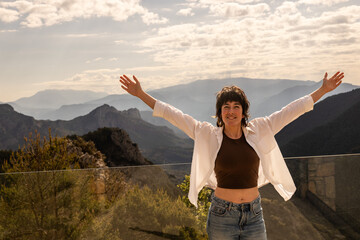 Woman stands with arms wide on mountain overlook. She wears a white jacket over a brown top and jeans. Distant mountains and partly cloudy sky frame the scene. Her smile shows joy and freedom © VICTOR