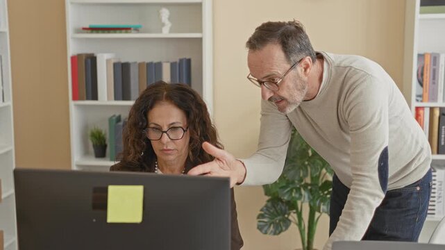 Woman at computer as man leans over pointing to monitor in a building office with bookshelves and a sticky note on the screen; teamwork focus.