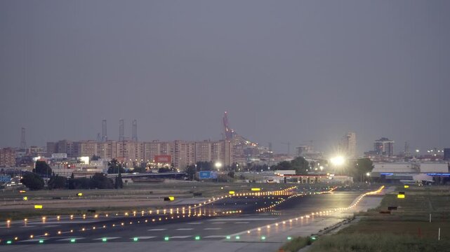 Aircraft is landing, airport runway edge and centerline lights glow at dusk, port cranes and city skyline silhouetted against a gray-purple sky in the background