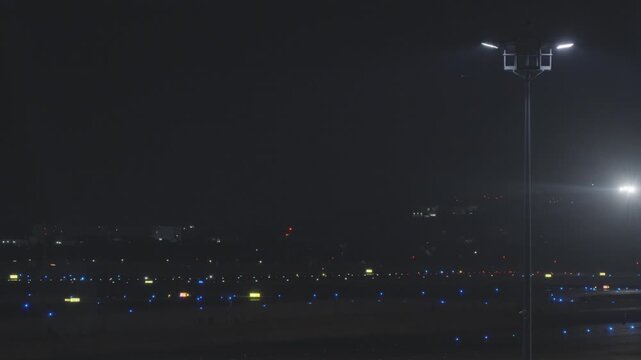 Dark airport apron at night with rows of blue taxiway edge lights and a tall floodlight mast, faint aircraft landing light visible at edge