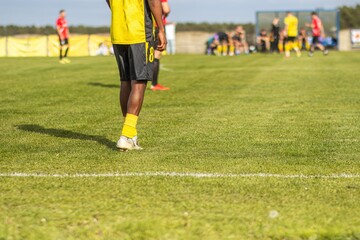 Obraz premium Male soccer player in yellow jersey and black shorts stands on green grass field, teammates and opposing players visible in the background during a match