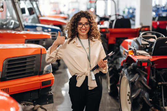 Female agricultural equipment sales representative stands in a showroom surrounded by orange tractors, holding a clipboard and giving a thumbs-up gesture