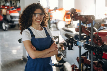 Female mechanic with curly hair wearing safety glasses and blue overalls stands confidently in a workshop surrounded by various vehicles and tools © standret