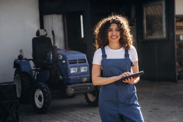 Female agricultural worker in blue overalls holding tablet stands outside a farm building with a blue tractor parked nearby on a sunny day © standret