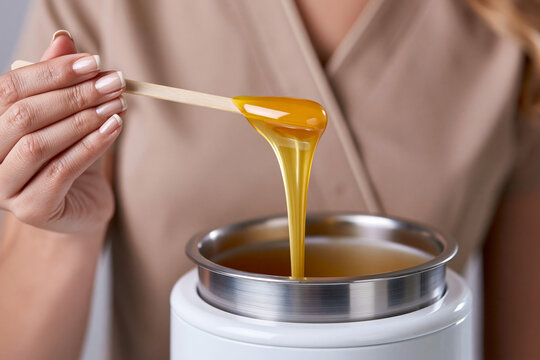 Professional beautician with French manicure holding a wooden spatula with dripping golden sugar paste over a heater. Sugaring hair removal process in a beauty salon for smooth skin and professiona...