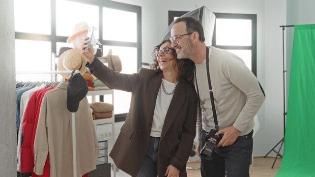 Woman wearing glasses holds smartphone and shows images while man photographer with camera and strap leans close in studio; collaborative joy.