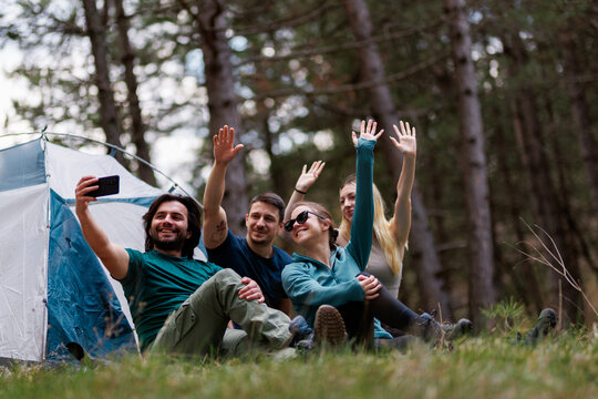 Friends enjoying a camping trip in the forest while taking a selfie together with smiles and waves