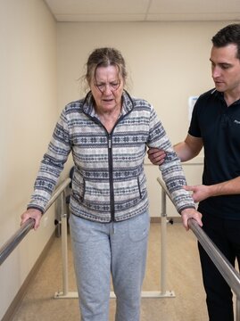 A woman with a determined expression walks between parallel bars, supported by a male physiotherapist. The scene captures the effort and support involved in rehabilitation.