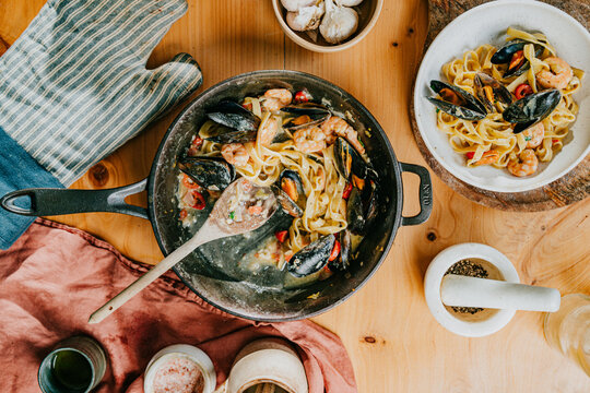 Sizzling pasta with mussels and shrimp in cast-iron pan. Wooden table set with garlic, herbs, and rustic napkins. Warm, inviting meal ready for family dinner or cozy night in. Great for food blogs