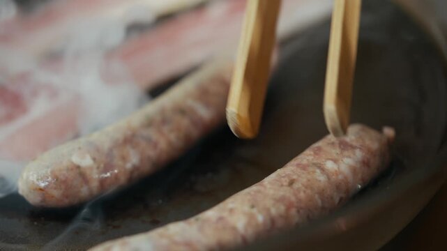 Sausages sizzle in hot pan, smoke curls upward. Wooden tongs gently flip sausages for even cooking. Steam rises as meat browns, aroma fills air. Ideal for food blogs, cooking tutorials