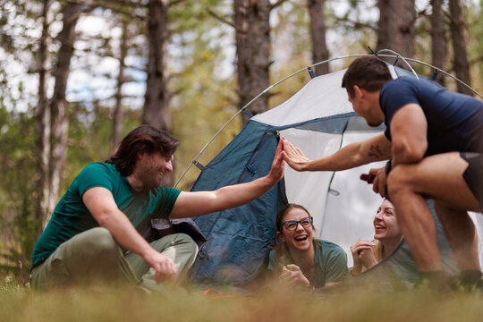 Friends enjoying a fun camping trip together in the forest, sharing laughter and high-fives under the trees during a sunny afternoon