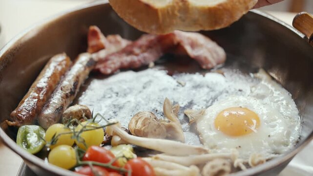 Hand places a slice of bread into a sizzling skillet. The pan holds sunny-side-up egg, sausages, bacon, mushrooms, and cherry tomatoes. Steam rises from hot food, indicating fresh cooking