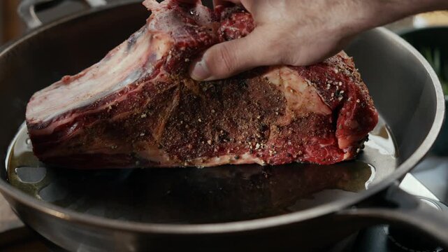 Hand places large, seasoned beef ribeye into hot skillet. Meat glistens with oil and spice. Steam rises as it hits the pan. Perfect for cooking tutorials or food blogs. High detail shows texture