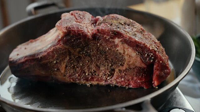 Large ribeye steak sizzles in hot pan. Seasoning spices coat meat surface. Steam rises as fat melts. Chef hand adjusts steak position. Perfectly seared crust forms on steak