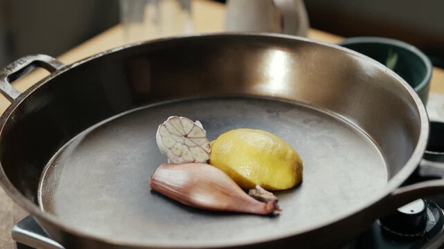 Pan holds garlic, lemon, and shallot on stovetop. Ingredients ready for cooking. Warm kitchen lighting enhances food preparation. Ideal for culinary tutorials or recipe videos