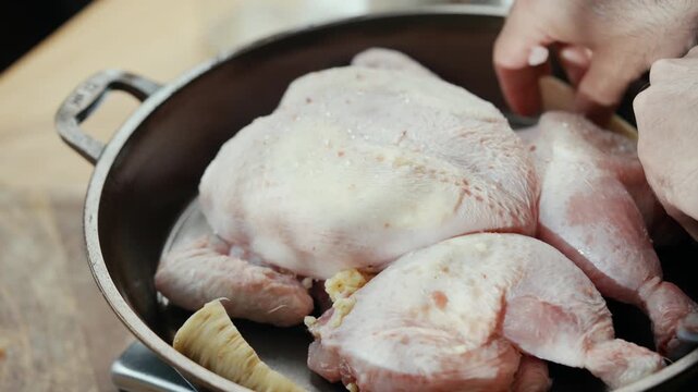 Raw chicken pieces rest in a metal pan on a wooden surface. Hand adds seasoning to the poultry. The action shows cooking preparation in detail. Ideal for recipes, food blogs, or culinary tutorials